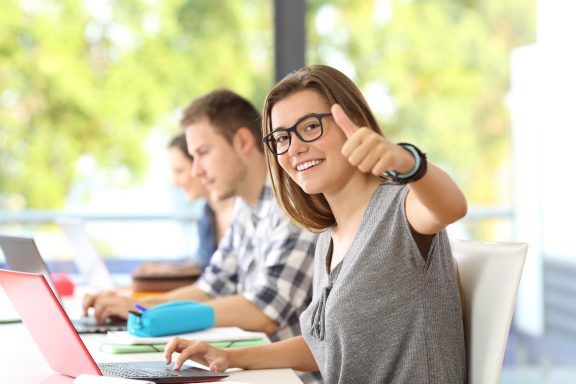 Junge Frau mit Brille gibt Daumen hoch, während sie am Laptop arbeitet.